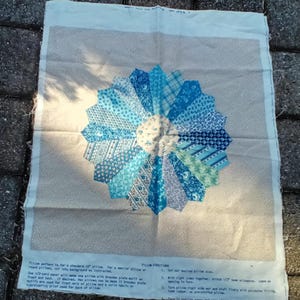 May include: A fabric panel featuring a Dresden plate design in shades of blue and white. The central motif resembles a flower, with instructions printed at the bottom. The panel is designed for a 16-inch pillow.