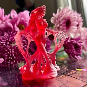 Könnte beinhalten: Eine durchscheinende rote Skulptur einer Figur mit ausgestreckten Armen, die einer Geisterform ähnelt. Die Skulptur steht vor einem Hintergrund aus rosa und lila Blumen, wodurch ein Kontrast aus Texturen und Farben entsteht.