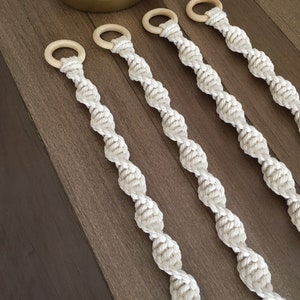 May include: Five white macrame plant hangers with wooden rings. The hangers are made of a thick, twisted rope and are designed to hang from the ceiling or a wall.