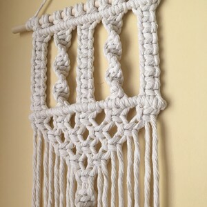 May include: Off-white macrame wall hanging with a geometric design. The piece features a wooden dowel at the top for hanging, with intricate knots and a fringe at the bottom. The wall decor is a neutral color.