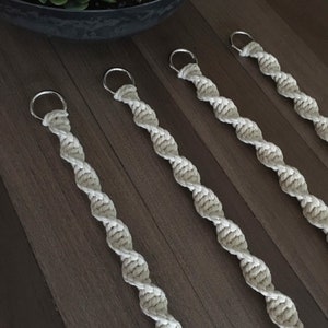 May include: Four macrame plant hangers with silver rings. The hangers are made of natural cotton rope and are hanging on a wooden surface.
