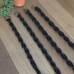May include: Five black macrame plant hangers with gold metal rings. The hangers are made of twisted rope and are hanging from a wooden surface.