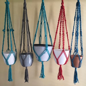 May include: Five macrame plant hangers in different colors, each with a pot. The hangers are made of cotton cord and have a variety of knot patterns. The pots are white, gray, and brown.