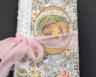 Könnte beinhalten: Ein handgefertigtes Notizbuch mit floralem Muster auf dem Einband und einer runden Illustration von zwei Kaninchen. Das Notizbuch ist mit einem rosa Band, Spitzenbesatz und dekorativen Knöpfen verziert. Das Design ist vintage und skurril.