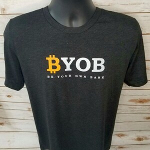 May include: Dark gray t-shirt with the text "BYOB" in yellow and white, with a Bitcoin symbol. Below the text is the phrase "BE YOUR OWN BANK". The shirt is displayed on a mannequin.