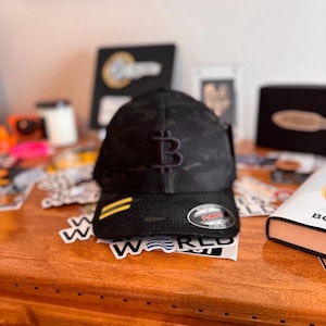 Puede incluir: Una gorra de béisbol negra de camuflaje con una franja amarilla y un símbolo de Bitcoin bordado en negro. La gorra está sobre una mesa de madera con varias pegatinas.