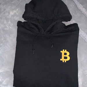 May include: Black hooded sweatshirt with a gold embroidered Bitcoin symbol on the front.