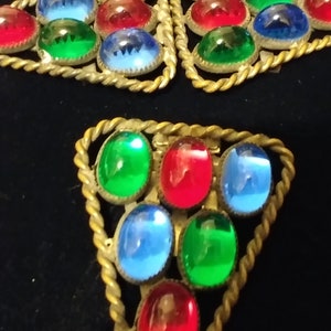 May include: Three gold-toned triangular brooches with a rope-like border. Each brooch is adorned with oval-shaped glass stones in shades of red, blue, and green.