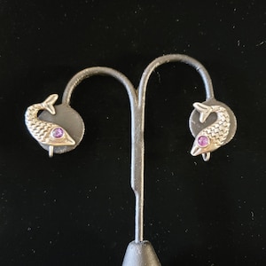May include: A pair of silver earrings shaped like fish with a purple gemstone in the center of each fish.