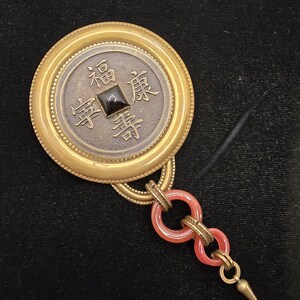 May include: A vintage coin-shaped pendant with gold-toned and silver-toned metal. The coin features Chinese characters and a black square stone. Attached are two red circular links and a gold-toned spike pendant.