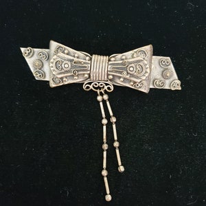 May include: A silver bow brooch with a dangling chain of beads. The bow is intricately detailed with floral patterns and swirls.