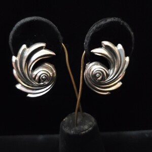 May include: A pair of silver-toned earrings featuring a swirling, shell-like design with layered, curved segments that spiral toward a central point. The metallic surface has a polished finish with dark recessed areas that highlight the intricate shape.