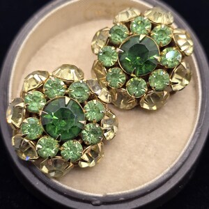 May include: A pair of vintage clip-on earrings. Each earring features a central, large, emerald-green stone surrounded by smaller green and yellow-green stones. The stones are set in a gold-tone metal setting, creating a floral design. The earrings are displayed in a gray jewelry box.