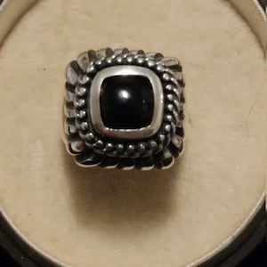 May include: A silver ring with a black square gemstone set in a decorative border.