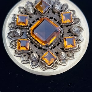 May include: A round, silver-toned brooch with a decorative filigree design. The brooch is adorned with eight amber-colored glass stones in a square shape.
