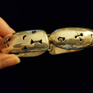 May include: Silver bracelet with a textured, oval-shaped design. The bracelet has cut-out shapes, including a crescent moon, a cross, and a triangle.