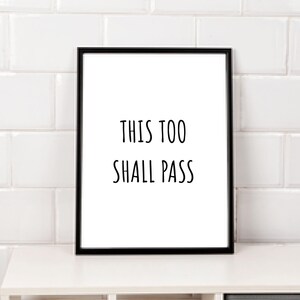 May include: A black framed print with the text "THIS TOO SHALL PASS" on a white background. The print is sitting on a white shelf with a gray drawer.