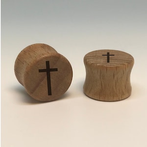 May include: Two wooden ear gauges with a cross design. The gauges are a light brown color and have a smooth finish.