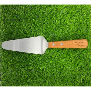 May include: A stainless steel cake server with a wooden handle. The handle is engraved with "Mr. & Mrs. Carson". The server is lying on a bed of green grass.