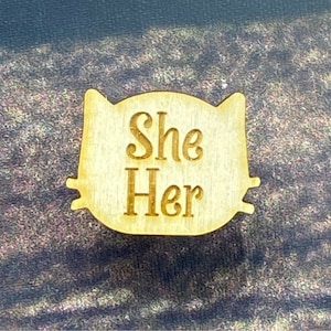May include: A wooden cat-shaped pin with the words "She Her" engraved in a serif font. The pin is light brown and has small, pointed ears and a rounded head. The background is a textured, dark surface.