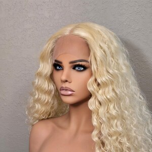 May include: A blonde, curly wig on a mannequin head. The wig features a lace front and long, wavy strands. The mannequin has blue eyes, dark eyebrows, and full lips. The background is a textured, light gray wall.