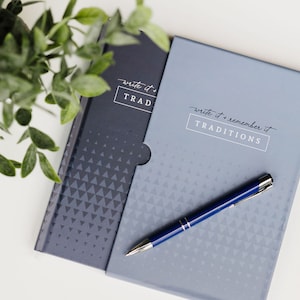 May include: Two blue journals with a geometric pattern on the cover. The top journal has the text "write it + TRADITIONS" on the cover. The bottom journal has the text "write it + remember it TRADITIONS" on the cover. A blue pen is lying on top of the journals.