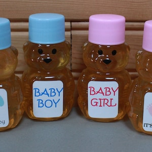 May include: Six bear-shaped honey bottles with blue and pink lids. The bottles have labels that say "Baby Boy," "It's a Boy," "Baby Girl," and "It's a Girl." The honey is amber-colored.