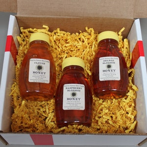 May include: A white box with red and white stripes on the sides. Inside the box, there are three glass bottles of honey. The bottles are filled with honey and have yellow lids. The labels on the bottles read "Clover Honey", "Orange Blossom Honey", and "Raspberry Blossom Honey".