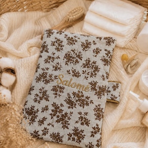May include: A rectangular fabric baby diaper clutch featuring a light blue background with a dark brown floral pattern. The name "Salom&eacute;" is embroidered in gold thread across the center. The clutch has a side tab closure and a quilted texture.