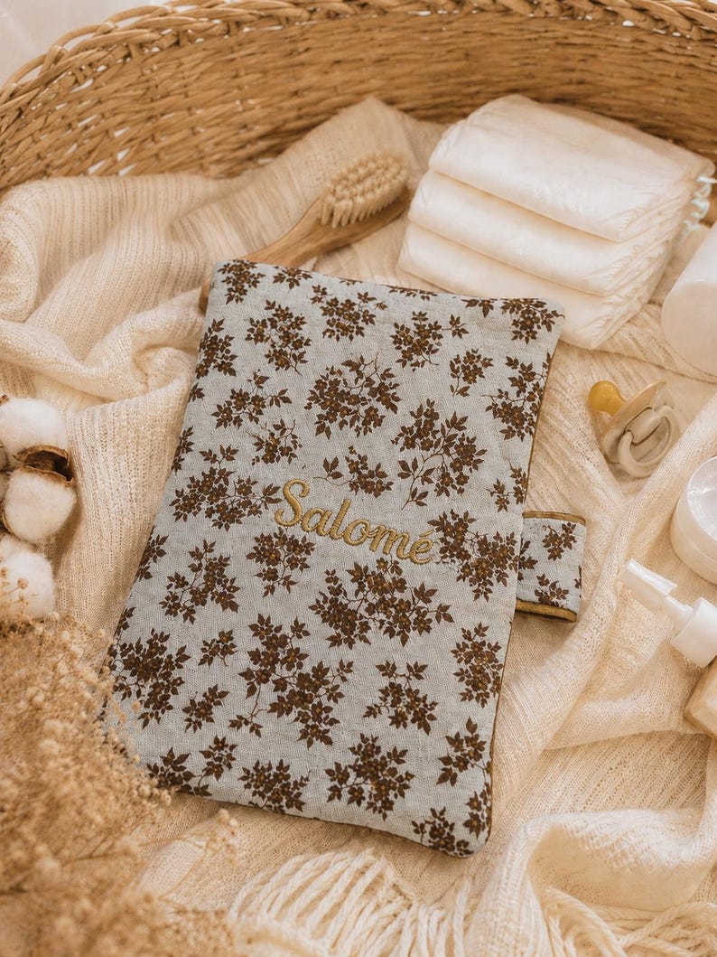 May include: A rectangular fabric baby diaper clutch featuring a light blue background with a dark brown floral pattern. The name "Salom&eacute;" is embroidered in gold thread across the center. The clutch has a side tab closure and a quilted texture.