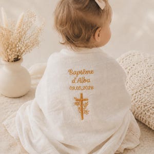 Peut inclure: Une couverture d'emmaillotage en mousseline blanche avec une broderie dorée indiquant Baptême d'Alba, la date 09.05.2026 et une croix ornée de branches. Le tissu présente une texture quadrillée et recouvre un enfant portant un petit nœud beige.