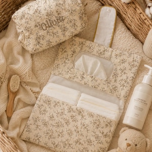 Peut inclure: Une station à langer pour bébé à imprimé floral beige avec une trousse de maquillage assortie brodée du nom "Olivia". La scène comprend une brosse, une lotion, des couches et un ours en peluche, le tout dans un panier en osier.