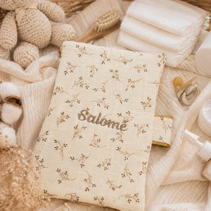May include: A rectangular fabric case featuring a cream background with a delicate brown floral pattern and the name Salom&eacute; embroidered in brown thread. The case has a small side tab and is positioned among various baby care items.