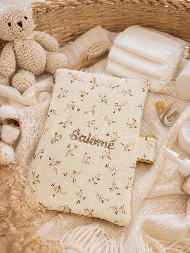 May include: A rectangular fabric case featuring a cream background with a delicate brown floral pattern and the name Salom&eacute; embroidered in brown thread. The case has a small side tab and is positioned among various baby care items.