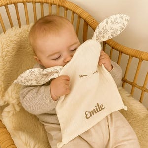 Peut inclure: Un bébé câline une couverture de sécurité crème avec des oreilles de lapin et le nom "Emile" brodé dessus. Les oreilles de lapin ont un motif floral. Le bébé porte un pull gris clair et un pantalon beige.
