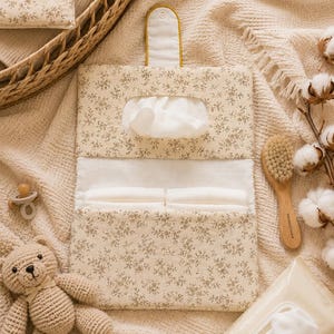 May include: A rectangular fabric diaper clutch featuring a beige floral pattern. The clutch has a top flap with a snap closure, an opening for dispensing wipes, and an interior pocket containing two folded white diapers.