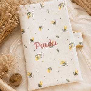 May include: A rectangular fabric cover featuring a cream background with a repeating pattern of small yellow flowers and green leaves. The name Paula is embroidered in pink thread in the center. A small tab extends from the right side of the cover.