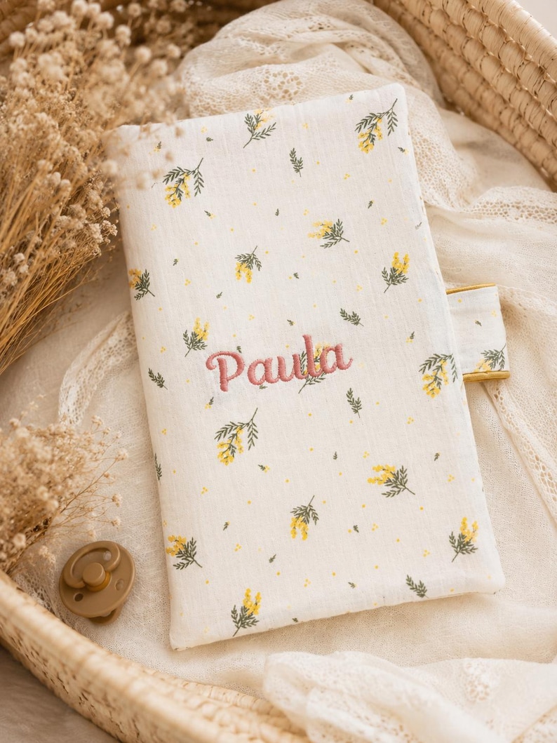 May include: A rectangular fabric cover featuring a cream background with a repeating pattern of small yellow flowers and green leaves. The name Paula is embroidered in pink thread in the center. A small tab extends from the right side of the cover.