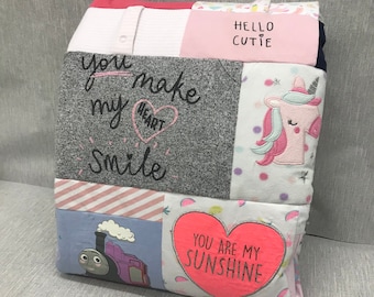 Puede incluir: Una colcha de retazos rosa y gris con varios dichos bordados, incluyendo "You make my heart smile", "Hello Cutie" y "You are my sunshine". La colcha también presenta un unicornio y un tren de dibujos animados.