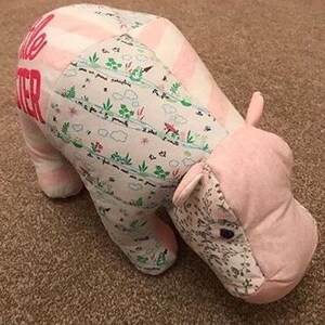 May include: A pink and white striped and floral patterned stuffed hippopotamus toy. The hippopotamus is lying on its side with its head resting on the ground. The hippopotamus has a pink nose and black eyes. The hippopotamus has the words "Little Sister" embroidered on its back.