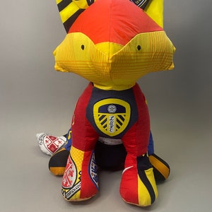 May include: A red, yellow, and black fox plush toy made from different pieces of fabric. The fox is wearing a blue and white soccer jersey with a crest that says "LUFC".