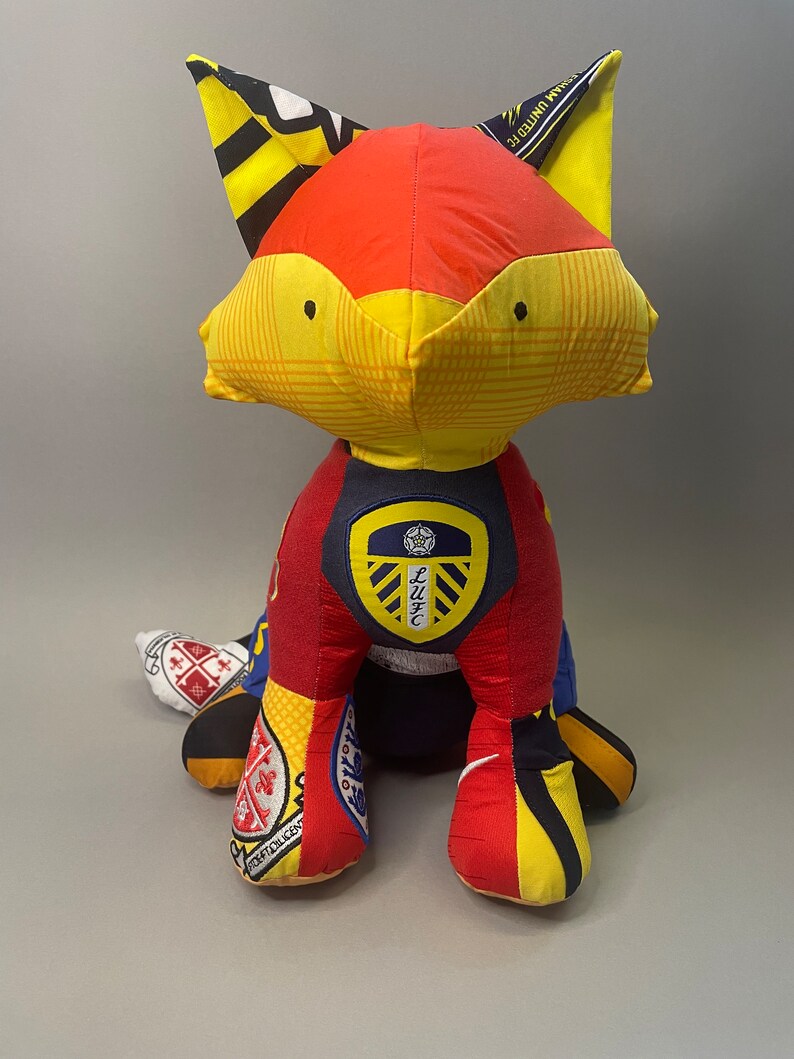 May include: A red, yellow, and black fox plush toy made from different pieces of fabric. The fox is wearing a blue and white soccer jersey with a crest that says "LUFC".