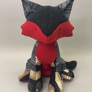 May include: A red and black fox plush toy with a patterned fabric design. The fox is sitting with its legs crossed and its tail curled behind it.