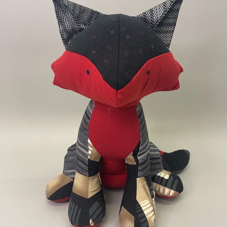 May include: A red and black fox plush toy with a patterned fabric design. The fox is sitting with its legs crossed and its tail curled behind it.