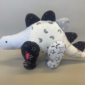 May include: A black and white patchwork dinosaur stuffed animal with a striped body and black and white star patterned legs. The dinosaur has a pink heart and the words "stay strong" embroidered on one leg.