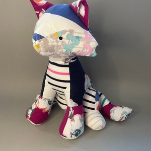 May include: A patchwork cat toy made from different fabrics, including floral prints, stripes, and solid colors. The cat is sitting with its legs outstretched and has a playful expression.