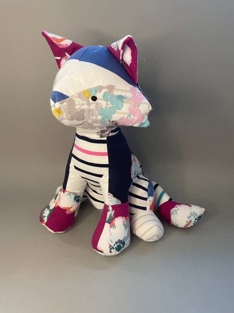 May include: A patchwork cat toy made from different fabrics, including floral prints, stripes, and solid colors. The cat is sitting with its legs outstretched and has a playful expression.
