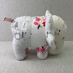 May include: A gray and white patchwork elephant plush toy with pink accents. The elephant has a floral pattern on one side and a pattern of animals and trees on the other side.