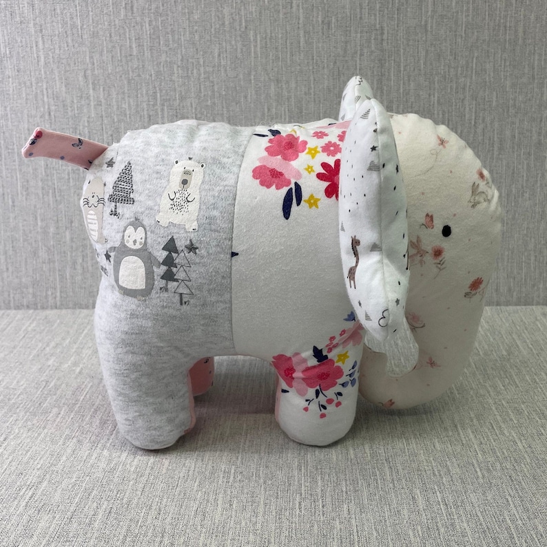 May include: A gray and white patchwork elephant plush toy with pink accents. The elephant has a floral pattern on one side and a pattern of animals and trees on the other side.