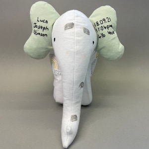 May include: A white stuffed elephant with green ears. The elephant has a rainbow design on its body and the text "Luca Joseph Stinson 18.09.21 11:04pm 4lb 4oz" is embroidered on the ears.